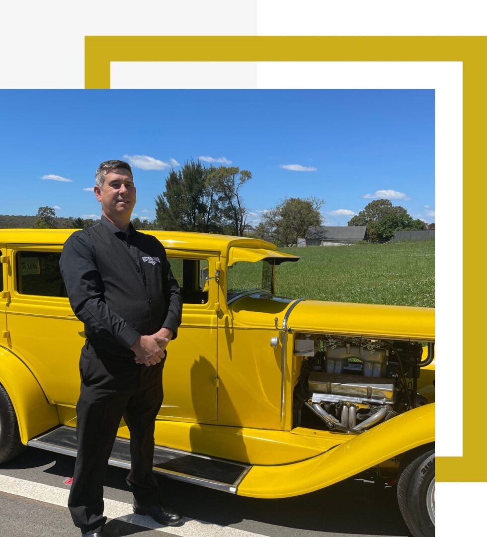 A professional chauffeur standing proudly by our yellow hot rod, offering top-notch service for vintage car hire Sydney