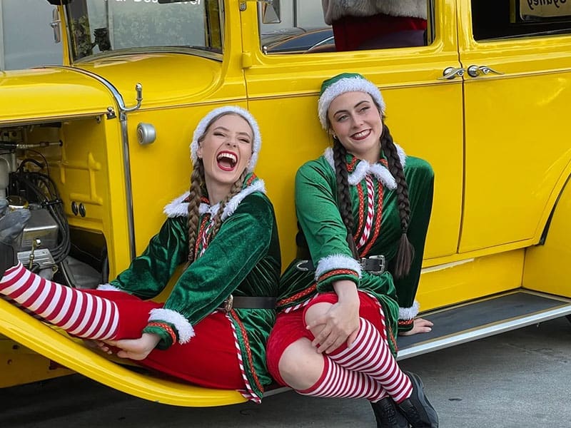 Santa's merry elves posing on the vibrant yellow hot rod, creating a festive scene for the vintage car photo shoot